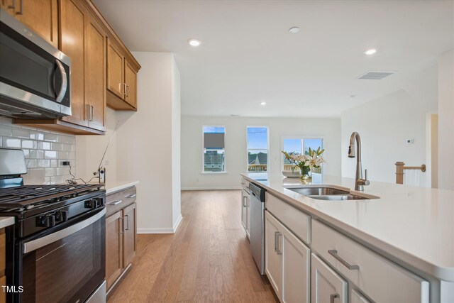 kitchen with sink, a kitchen island with sink, stainless steel appliances, light hardwood / wood-style floors, and decorative backsplash