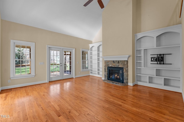 unfurnished living room featuring ceiling fan, french doors, a stone fireplace, built in features, and light hardwood / wood-style flooring