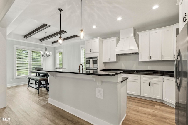 kitchen featuring white cabinets, an island with sink, beam ceiling, decorative light fixtures, and premium range hood