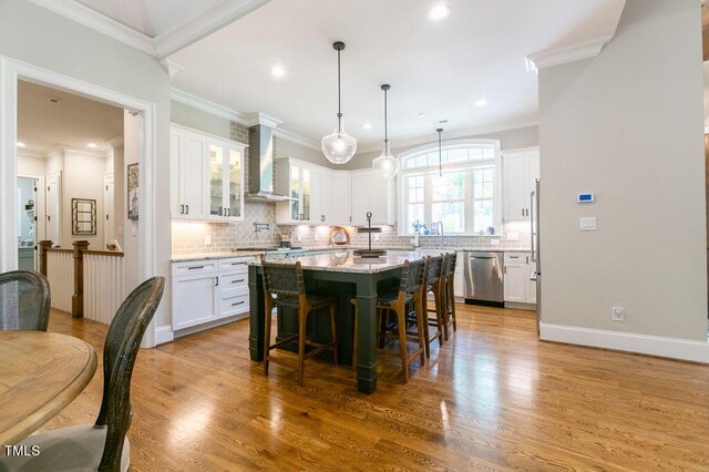 kitchen with a kitchen island, wall chimney range hood, wood-type flooring, stainless steel dishwasher, and white cabinets