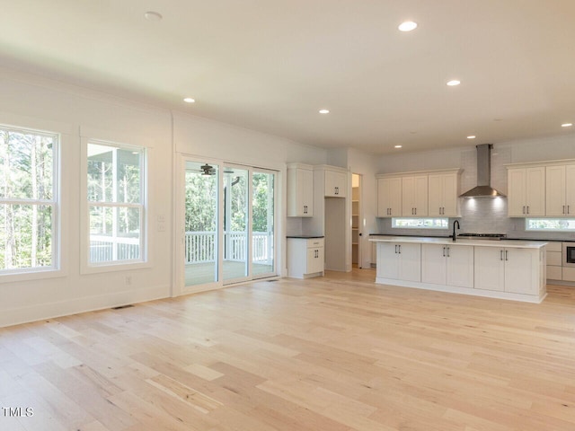 kitchen with light hardwood / wood-style floors, tasteful backsplash, wall chimney exhaust hood, a kitchen island with sink, and white cabinets