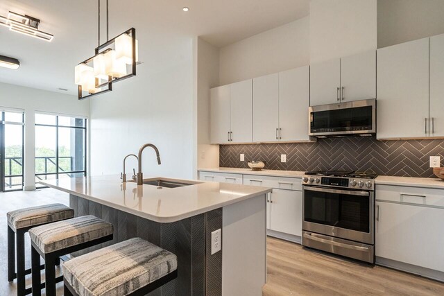 kitchen with appliances with stainless steel finishes, a kitchen island with sink, sink, decorative light fixtures, and light hardwood / wood-style flooring