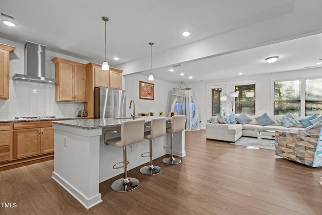 kitchen featuring light stone countertops, wall chimney exhaust hood, hanging light fixtures, a kitchen island with sink, and light wood-type flooring