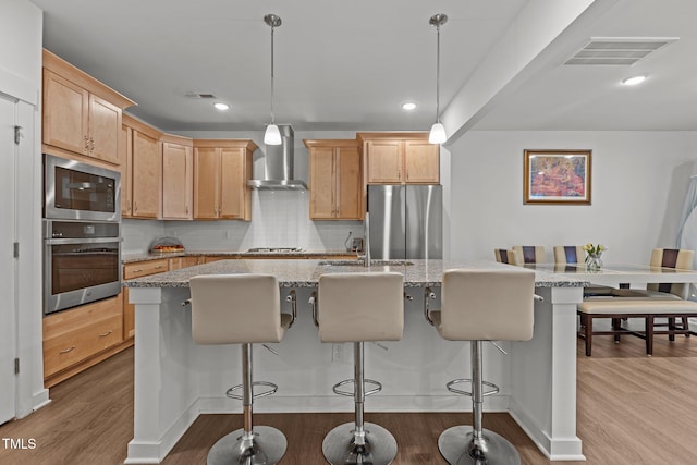 kitchen with stainless steel appliances, wall chimney exhaust hood, a center island with sink, and decorative light fixtures