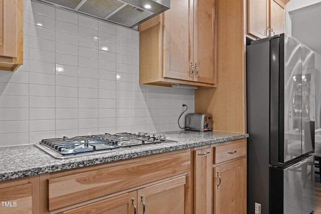 kitchen featuring stainless steel appliances, decorative backsplash, wall chimney exhaust hood, light stone countertops, and light brown cabinetry