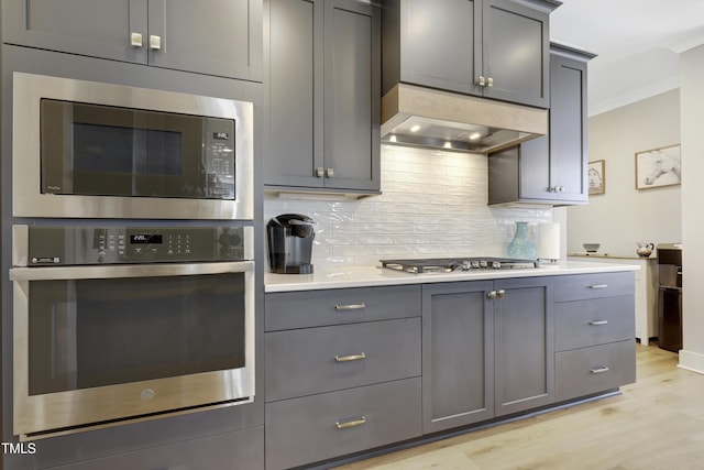 kitchen with exhaust hood, gray cabinets, light wood-type flooring, tasteful backsplash, and stainless steel appliances