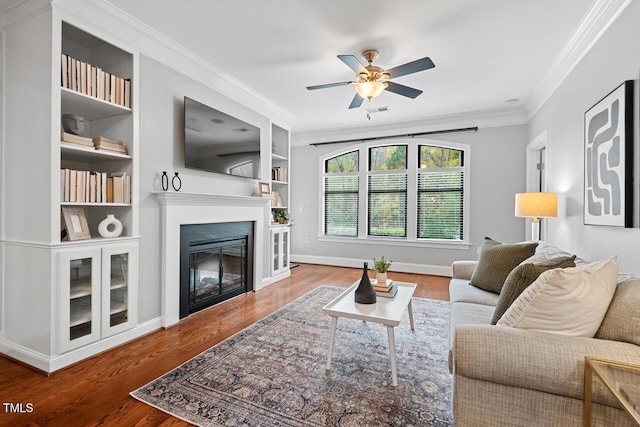 living area with ornamental molding, ceiling fan, wood finished floors, a glass covered fireplace, and built in features