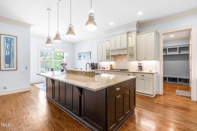 kitchen featuring wood-type flooring, light stone counters, crown molding, a kitchen breakfast bar, and a kitchen island with sink