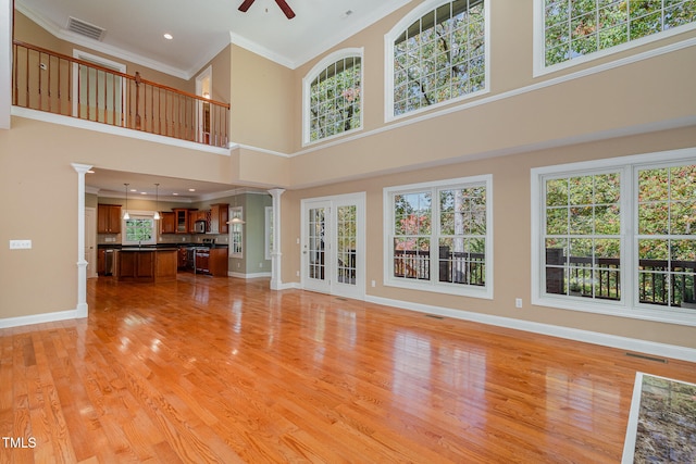 unfurnished living room with ceiling fan, a towering ceiling, crown molding, ornate columns, and light wood-type flooring