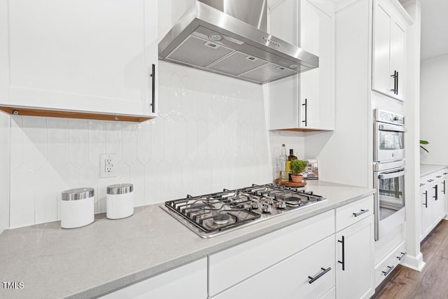 kitchen featuring wall chimney range hood, appliances with stainless steel finishes, decorative backsplash, white cabinets, and hardwood / wood-style flooring