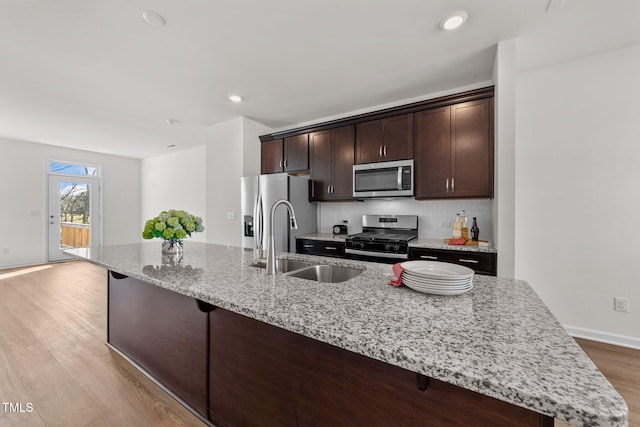 kitchen featuring dark brown cabinetry, an island with sink, light stone countertops, light wood-type flooring, and appliances with stainless steel finishes