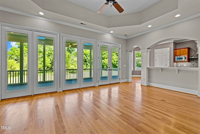 unfurnished living room featuring ceiling fan, a tray ceiling, crown molding, decorative columns, and light wood-type flooring