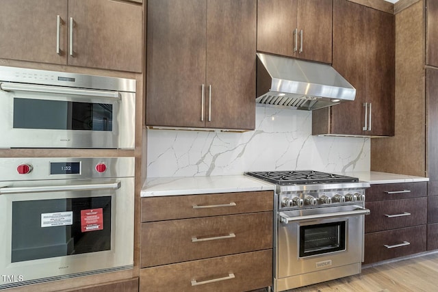 kitchen with appliances with stainless steel finishes, range hood, light stone counters, light hardwood / wood-style floors, and decorative backsplash