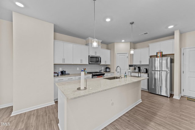 kitchen with appliances with stainless steel finishes, hanging light fixtures, light hardwood / wood-style floors, white cabinets, and a kitchen island with sink