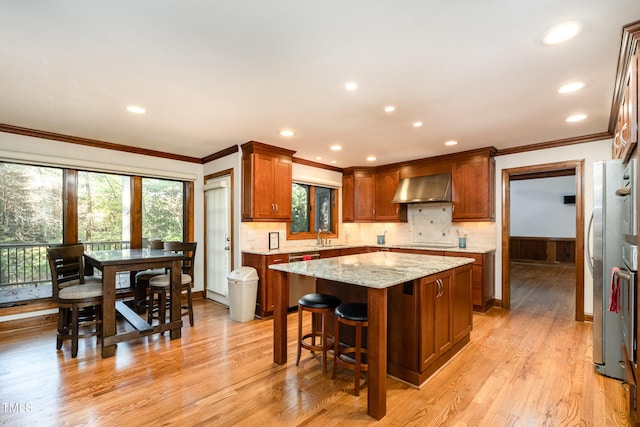 kitchen featuring ornamental molding, wall chimney range hood, a center island, light wood-type flooring, and light stone countertops