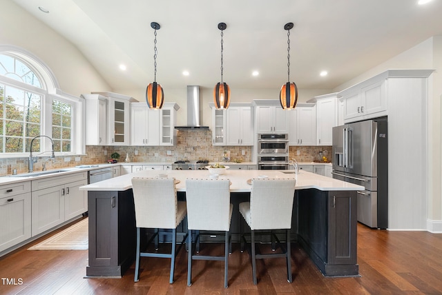 kitchen with pendant lighting, dark wood-type flooring, a center island with sink, wall chimney exhaust hood, and stainless steel appliances