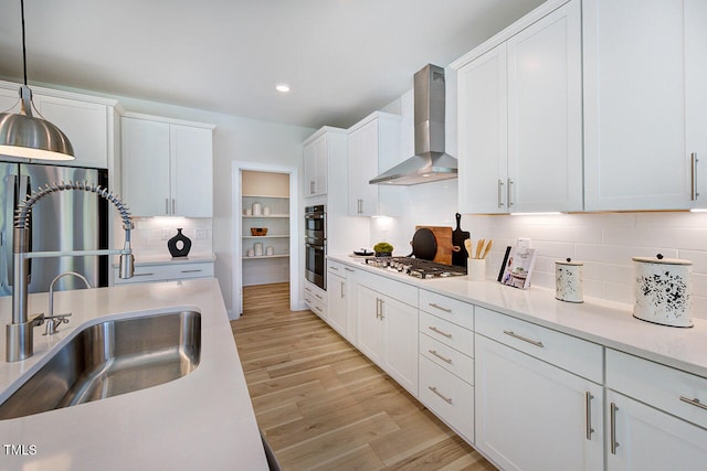 kitchen featuring sink, hanging light fixtures, stainless steel appliances, wall chimney range hood, and light wood-type flooring