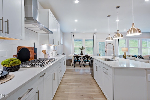 kitchen with light wood-type flooring, a kitchen island with sink, sink, wall chimney range hood, and white cabinetry