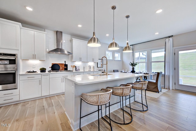 kitchen featuring appliances with stainless steel finishes, wall chimney exhaust hood, sink, a center island with sink, and white cabinetry