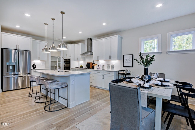 kitchen with stainless steel appliances, wall chimney range hood, pendant lighting, a center island with sink, and white cabinetry