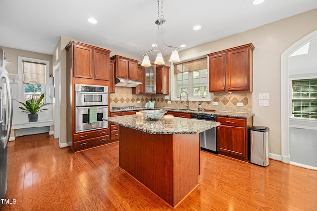 kitchen with light stone countertops, appliances with stainless steel finishes, light wood-type flooring, decorative light fixtures, and a kitchen island