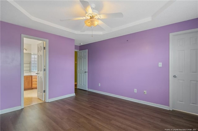 unfurnished bedroom with ensuite bathroom, a tray ceiling, dark hardwood / wood-style floors, and ceiling fan