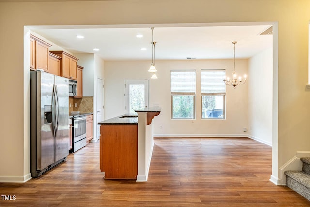 kitchen featuring hardwood / wood-style flooring, an island with sink, appliances with stainless steel finishes, tasteful backsplash, and decorative light fixtures