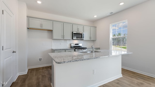 kitchen with light stone counters, an island with sink, stainless steel appliances, and hardwood / wood-style flooring