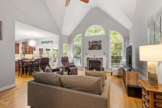 living room featuring french doors, light wood-type flooring, ceiling fan, high vaulted ceiling, and a stone fireplace