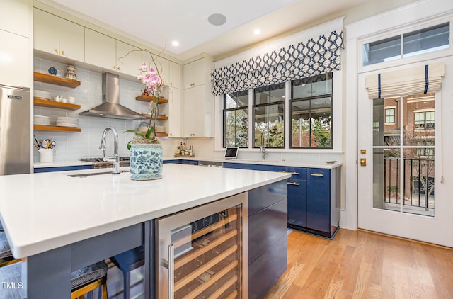 kitchen with light wood-type flooring, ornamental molding, wall chimney exhaust hood, wine cooler, and an island with sink