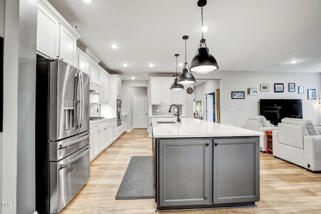 kitchen with light wood-type flooring, stainless steel appliances, a kitchen island with sink, decorative light fixtures, and white cabinetry