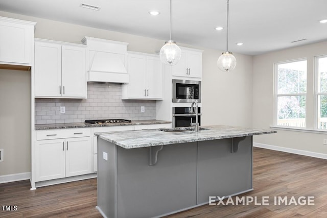 kitchen featuring decorative light fixtures, white cabinets, a kitchen island with sink, light stone countertops, and custom range hood