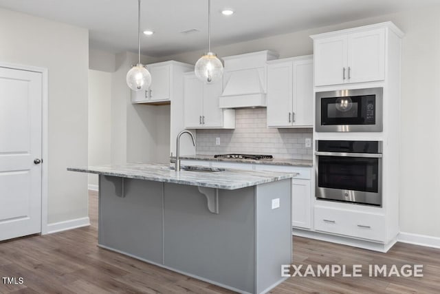 kitchen featuring sink, custom exhaust hood, decorative light fixtures, appliances with stainless steel finishes, and a kitchen island with sink