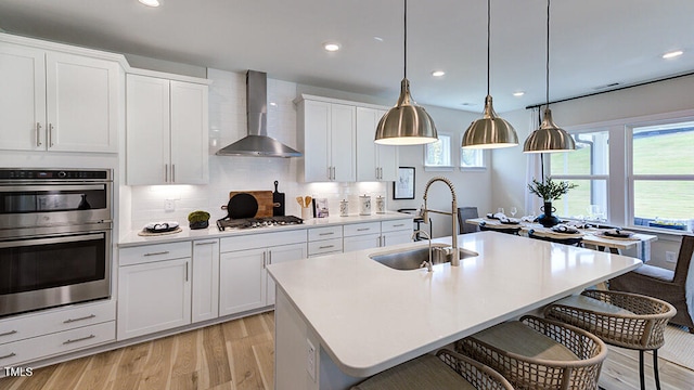 kitchen with appliances with stainless steel finishes, a kitchen island with sink, wall chimney range hood, white cabinetry, and hanging light fixtures