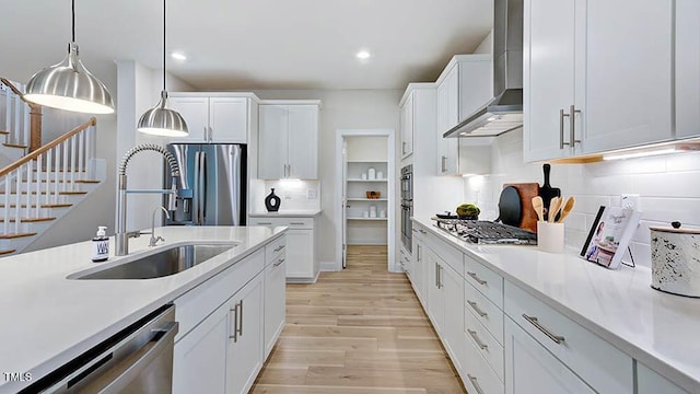 kitchen with pendant lighting, white cabinets, wall chimney exhaust hood, appliances with stainless steel finishes, and light hardwood / wood-style floors