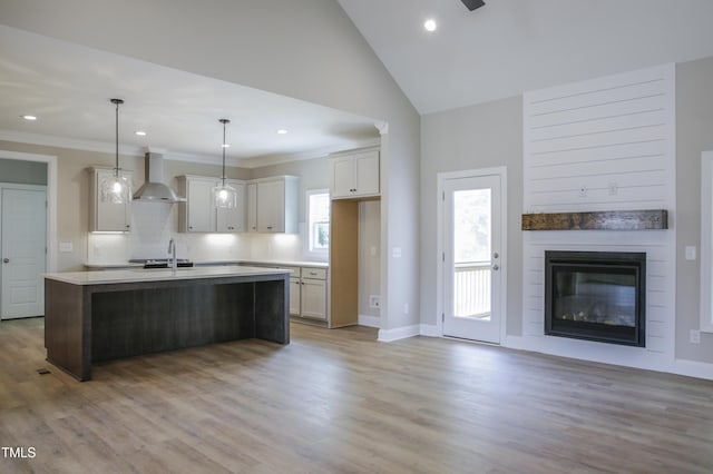 kitchen with wall chimney exhaust hood, pendant lighting, light hardwood / wood-style floors, white cabinetry, and an island with sink