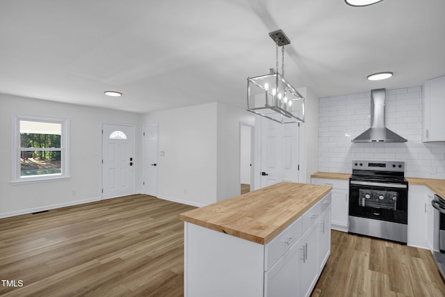 kitchen with pendant lighting, stainless steel electric range, white cabinets, wall chimney exhaust hood, and butcher block countertops