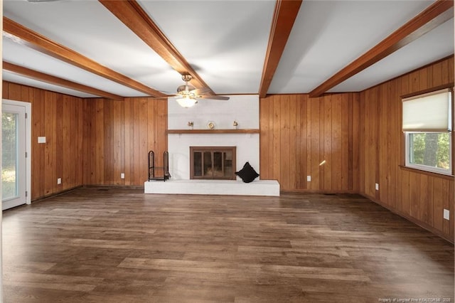 unfurnished living room with wooden walls, a ceiling fan, a brick fireplace, beamed ceiling, and dark wood finished floors