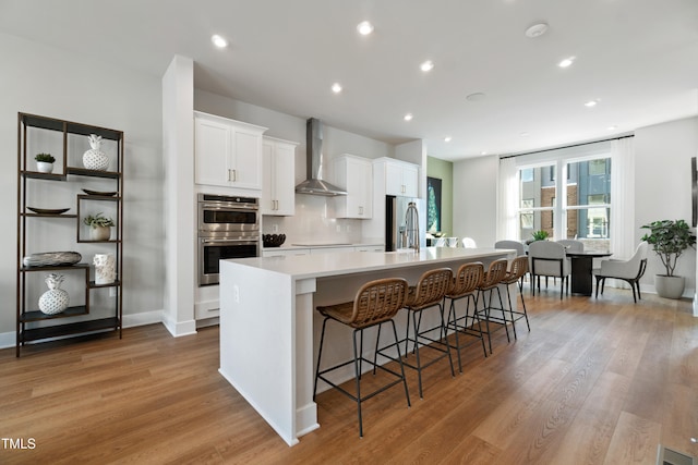 kitchen featuring wall chimney exhaust hood, a spacious island, light wood-type flooring, white cabinets, and appliances with stainless steel finishes