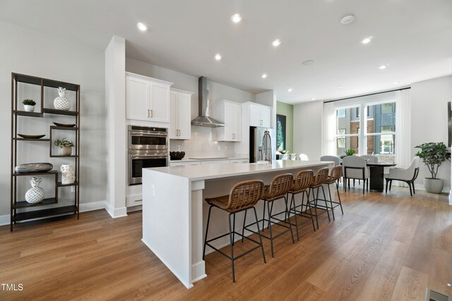 kitchen with light wood-type flooring, wall chimney exhaust hood, stainless steel appliances, a large island with sink, and white cabinets