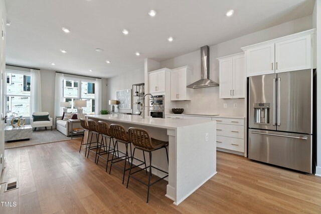 kitchen with appliances with stainless steel finishes, wall chimney exhaust hood, a breakfast bar, white cabinets, and an island with sink