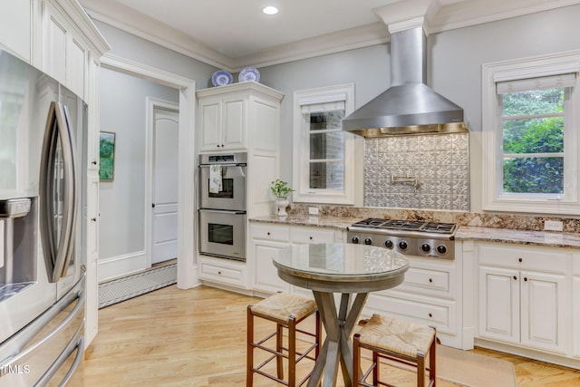 kitchen featuring wall chimney range hood, stainless steel appliances, white cabinets, and light stone countertops