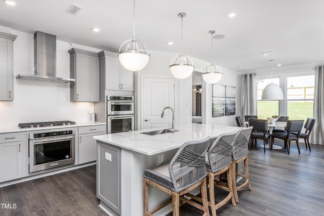 kitchen with gray cabinetry, wall chimney exhaust hood, stainless steel appliances, sink, and a center island with sink