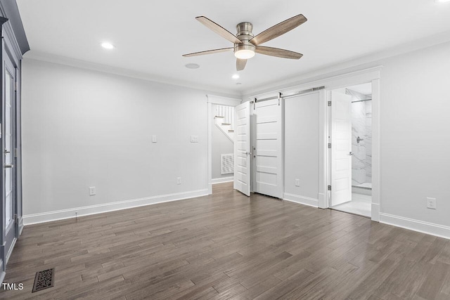 unfurnished bedroom featuring crown molding, ensuite bath, dark hardwood / wood-style floors, ceiling fan, and a barn door