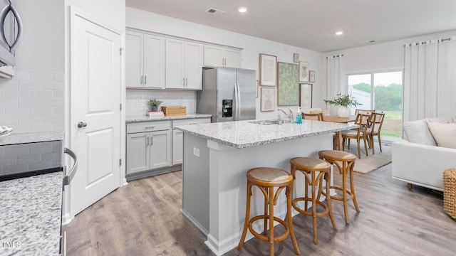 kitchen with light stone countertops, backsplash, a center island with sink, light hardwood / wood-style flooring, and stainless steel fridge with ice dispenser