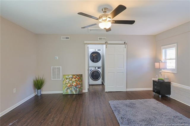 laundry area featuring ceiling fan, a barn door, stacked washer and dryer, and dark hardwood / wood-style flooring