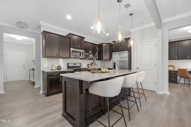 kitchen featuring light wood-type flooring, dark brown cabinets, stainless steel appliances, pendant lighting, and a center island with sink