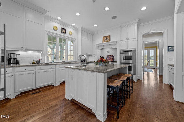 kitchen with light stone counters, a center island with sink, decorative backsplash, stainless steel double oven, and white cabinets