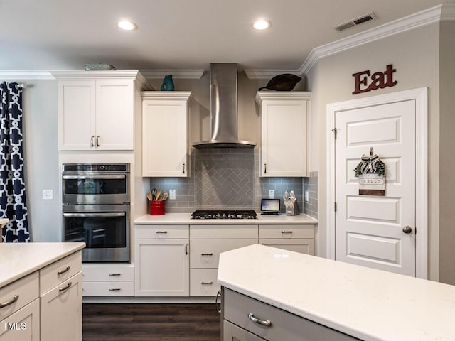 kitchen with dark wood-type flooring, white cabinets, wall chimney range hood, ornamental molding, and appliances with stainless steel finishes