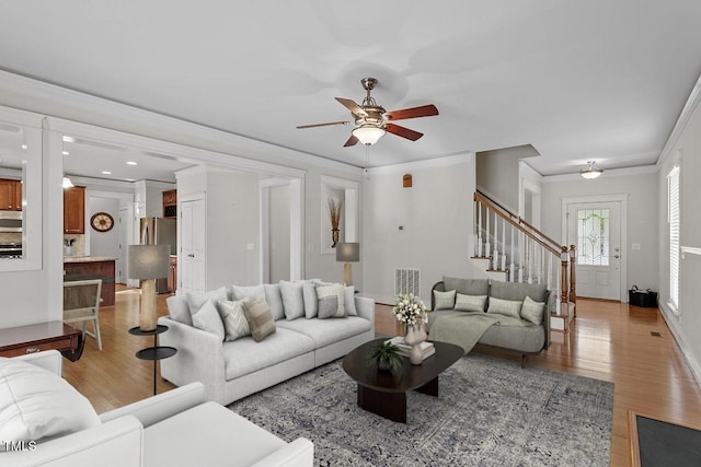 living room featuring crown molding, visible vents, light wood-style flooring, ceiling fan, and stairs